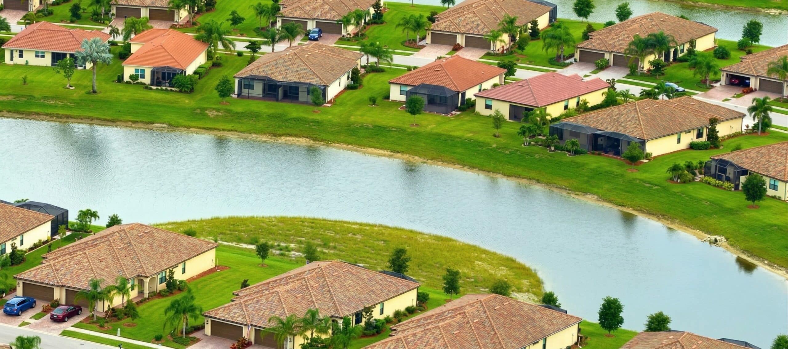 Aerial view of suburban houses with tile roofs arranged around a curved waterway, surrounded by green lawns and scattered palm trees, highlighting the importance of proper pond management for effective stormwater infrastructure.