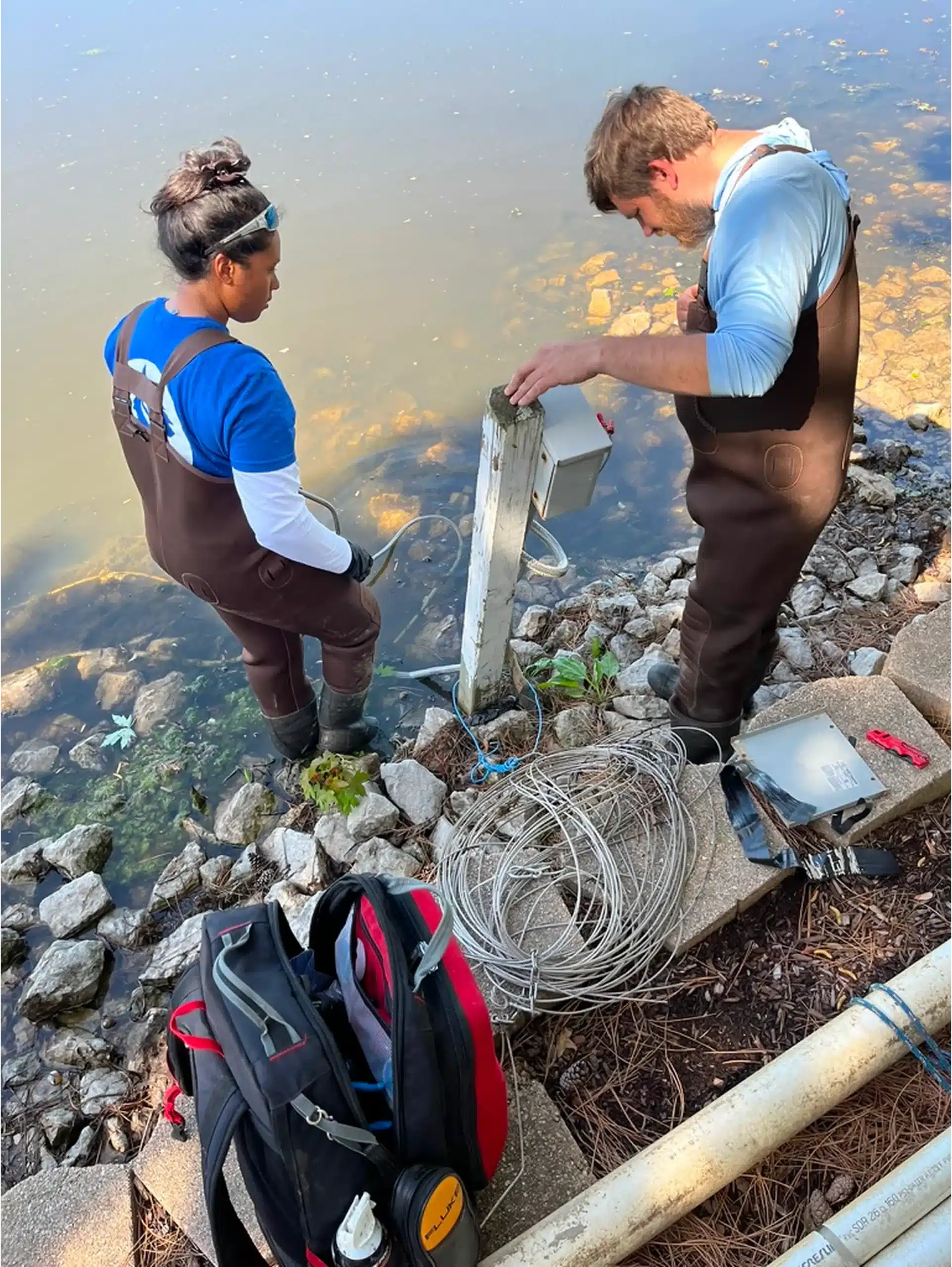 Two people wearing waders work on a water monitoring station by a rocky shoreline, with equipment and tools laid out nearby, supporting stormwater management efforts.