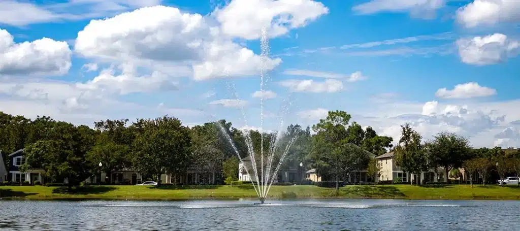 A water fountain sprays upward in the center of a pond, part of effective stormwater management, with houses and trees lining the grassy shore under a partly cloudy sky.