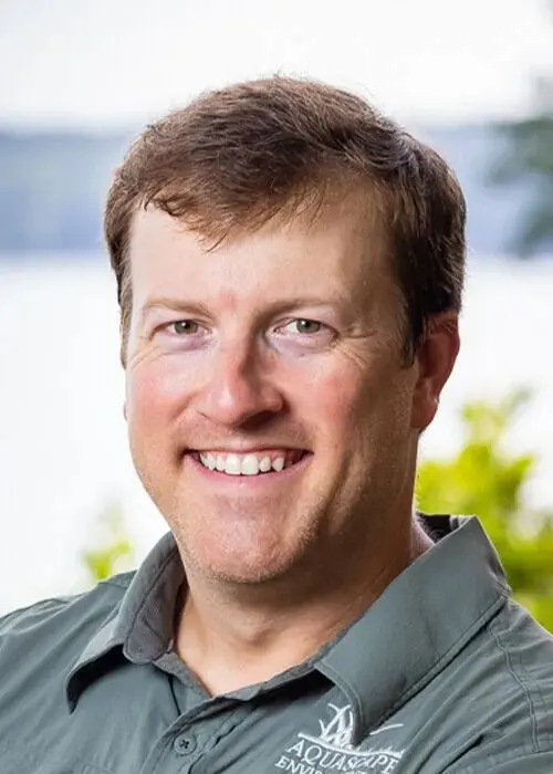 A man with short brown hair smiles at the camera, wearing a gray collared shirt with an "Aquascape" logo, outdoors with greenery and water in the background, reflecting his expertise in pond management.