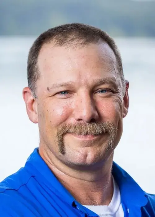 A man with short brown hair, a mustache, and blue eyes smiles at the camera. He is wearing a blue collared shirt, perfect for a day of pond management. The background is softly blurred.