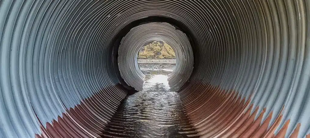View from inside a corrugated metal drainage pipe, used in stormwater management, with shallow water on the bottom, looking out toward a rocky landscape and daylight.