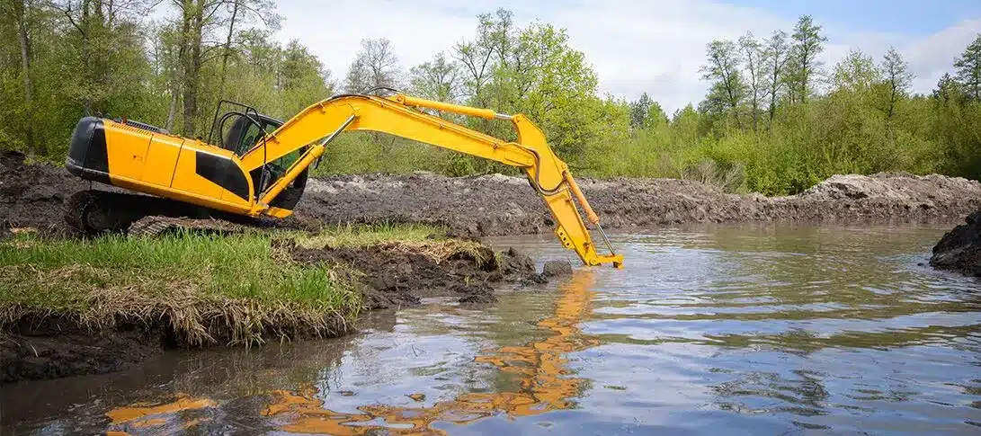 A yellow excavator is positioned on muddy ground, extending its arm into a body of water surrounded by trees and vegetation, showcasing essential pond maintenance.