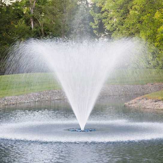 A fountain sprays water upward in a symmetrical pattern in the center of a small pond, surrounded by grass and trees.