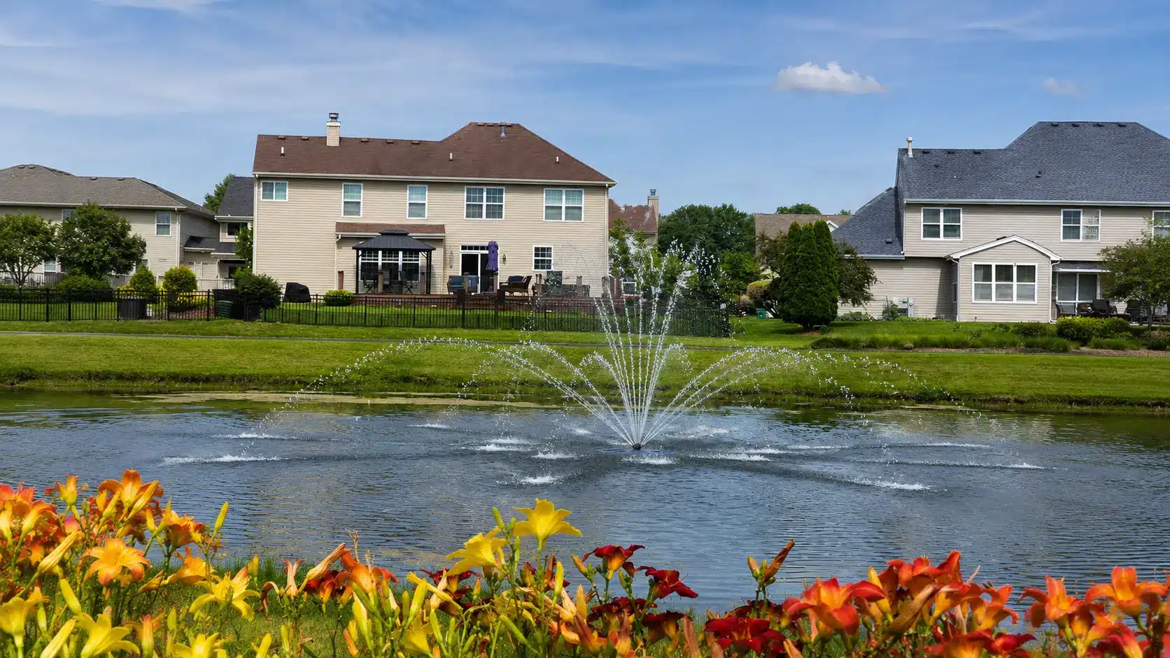 A pond with a water fountain and aeration systems in front of suburban houses, surrounded by green lawns and orange and yellow flowers under a blue sky.