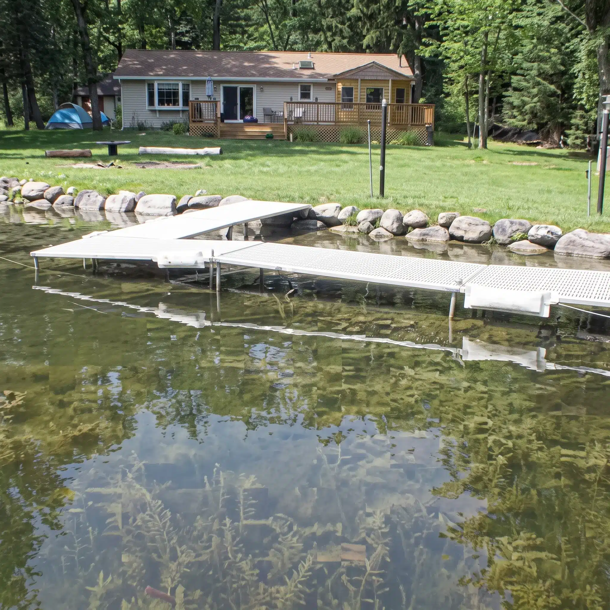 A white dock extends into a clear lake in front of a tan house with a wooden deck, surrounded by trees and green grass.