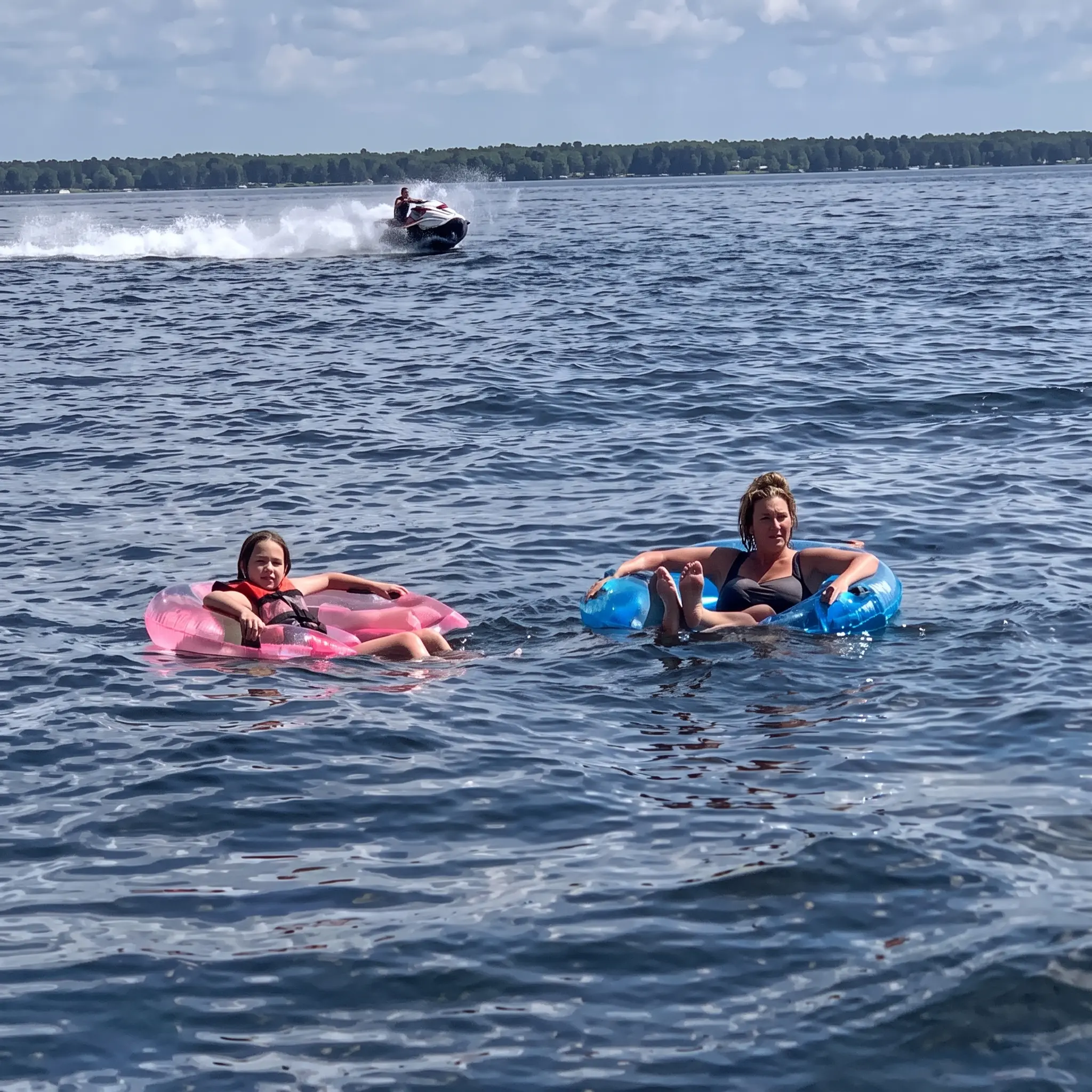 Two people float on inflatable rings in a lake, one pink and one blue. A jet ski speeds by in the background with trees visible on the shore under a partly cloudy sky.