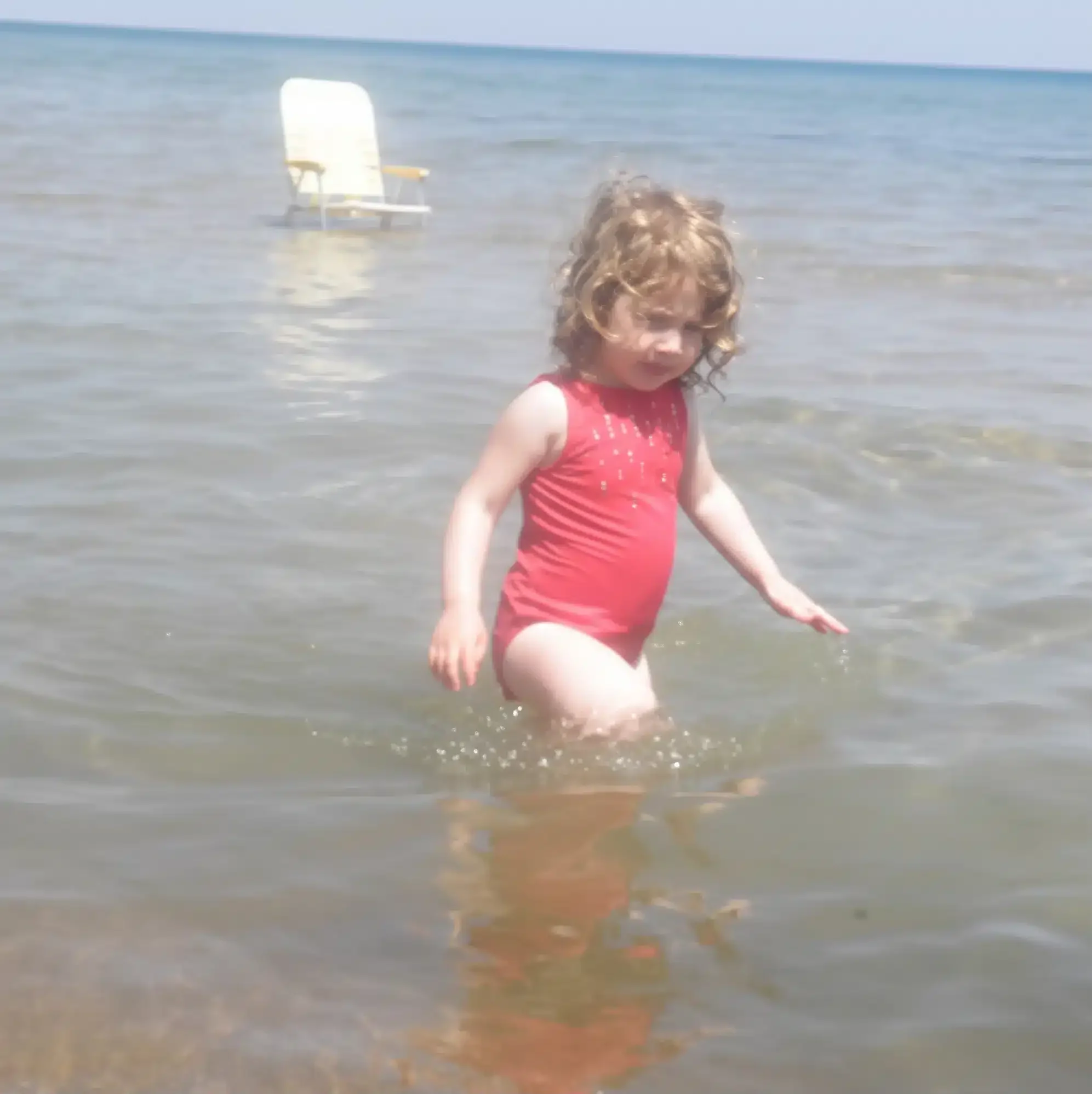 A young child in a red swimsuit stands in shallow water at the beach, with an empty chair in the background.