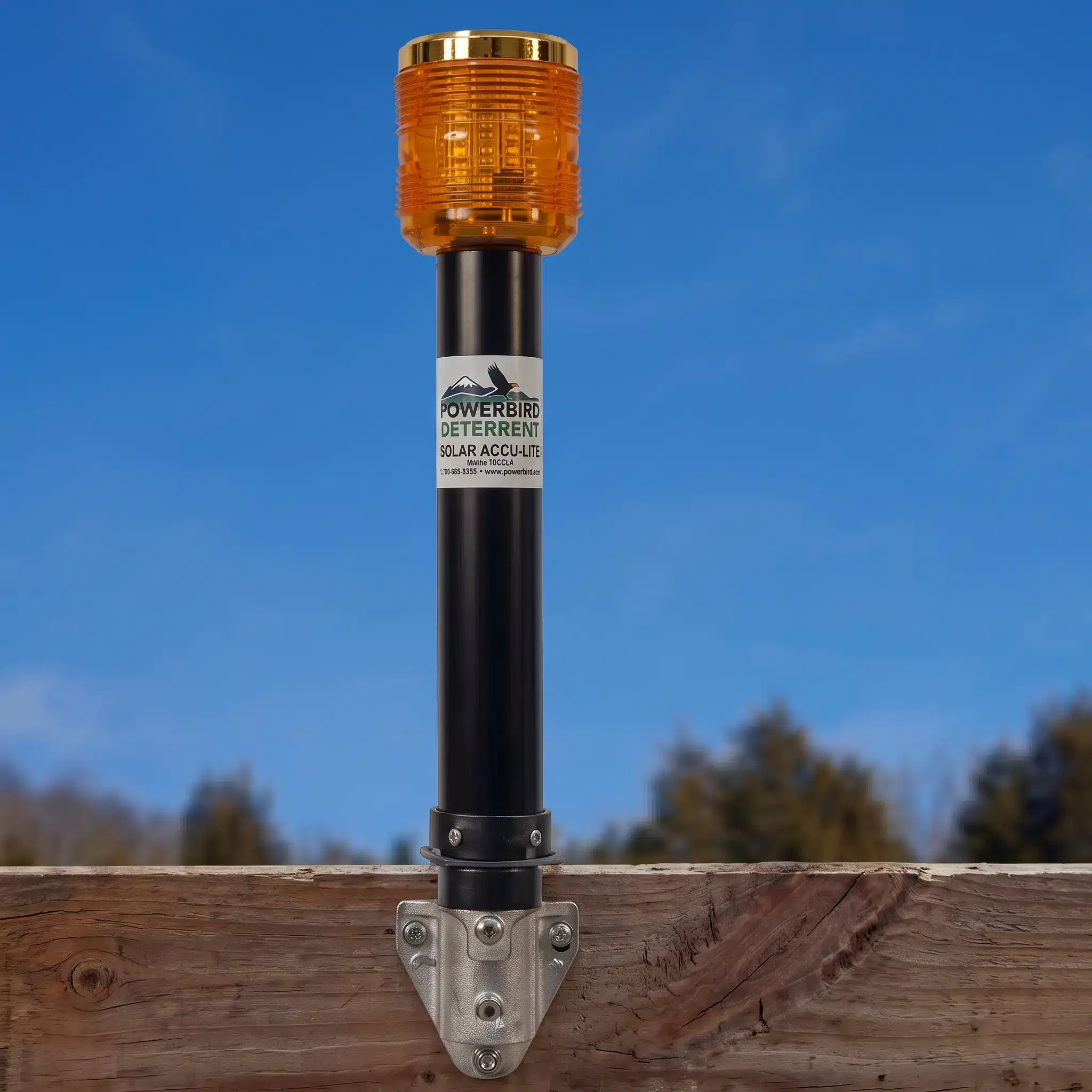Solar-powered bird deterrent device with an amber warning light mounted on a wooden surface outdoors, with trees and blue sky in the background.