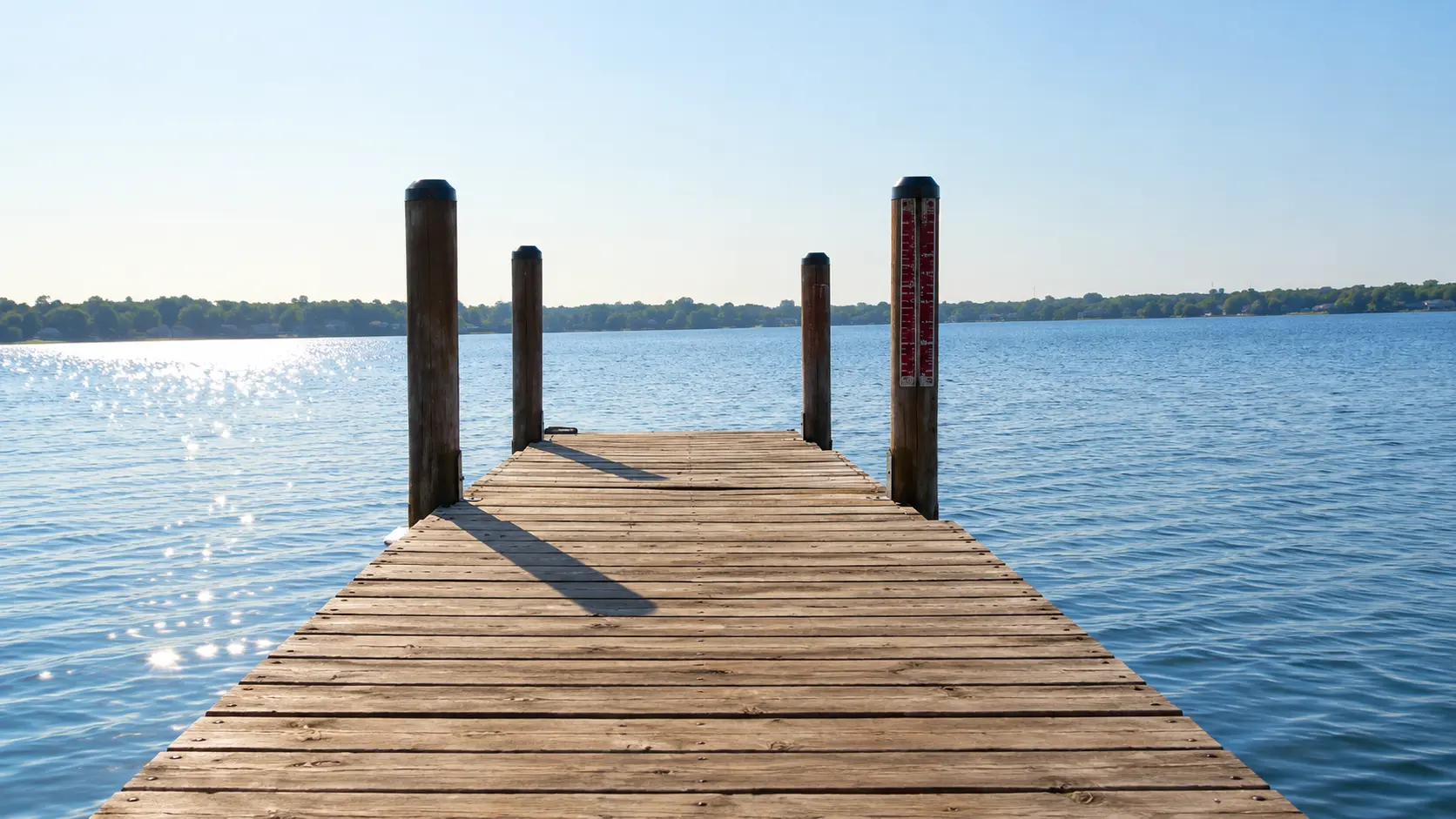 A wooden dock extends over calm lake water under a clear blue sky, with trees lining the distant shore.