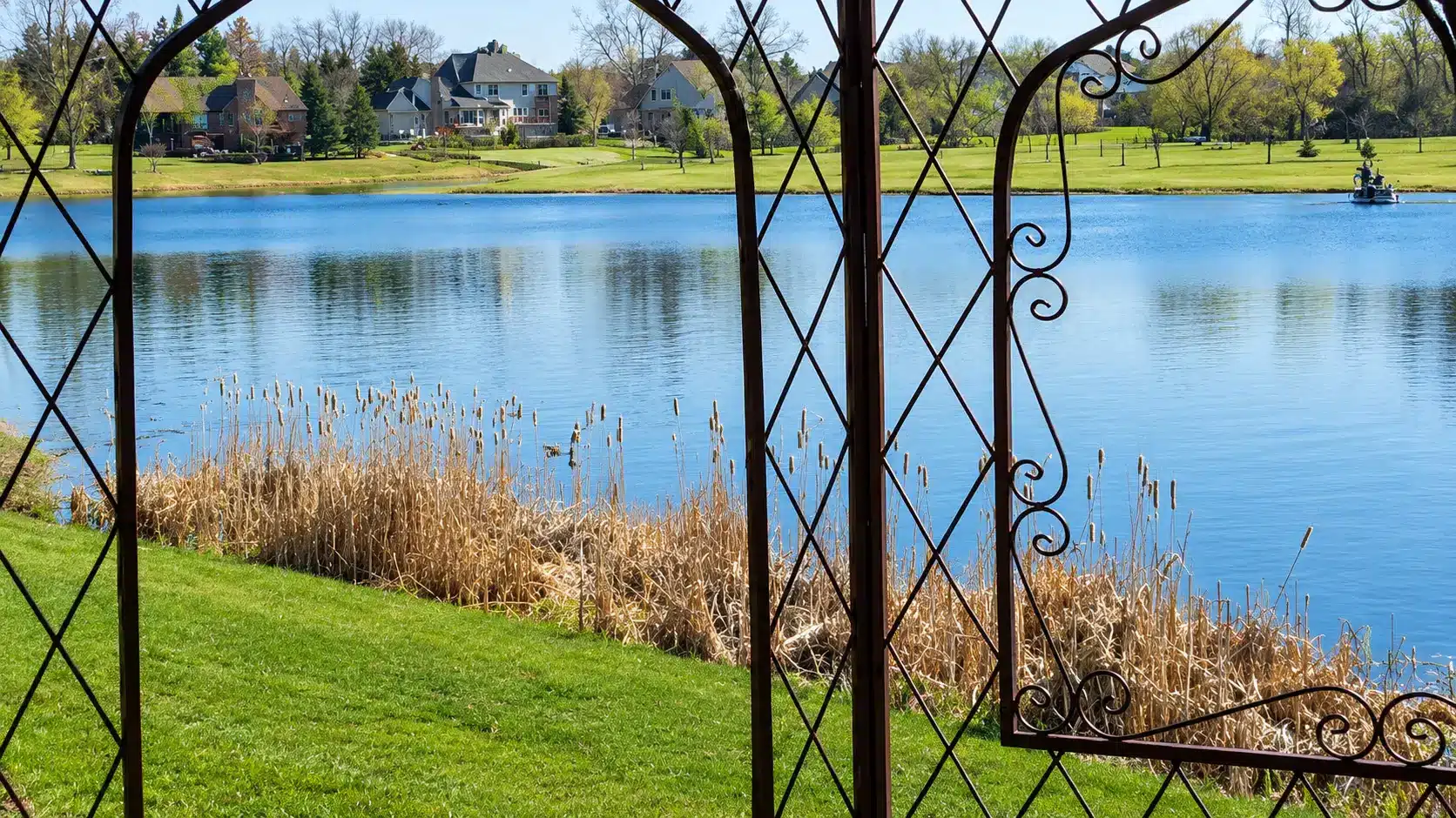 View of a calm lake bordered by reeds and grass, seen through an ornate metal garden structure, with houses and trees in the background.