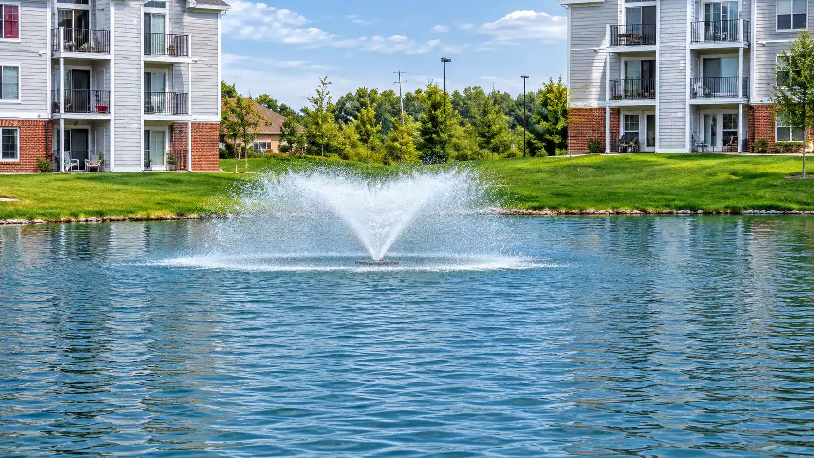 A water fountain sprays in the center of a pond, surrounded by grassy areas and modern apartment buildings under a partly cloudy sky.