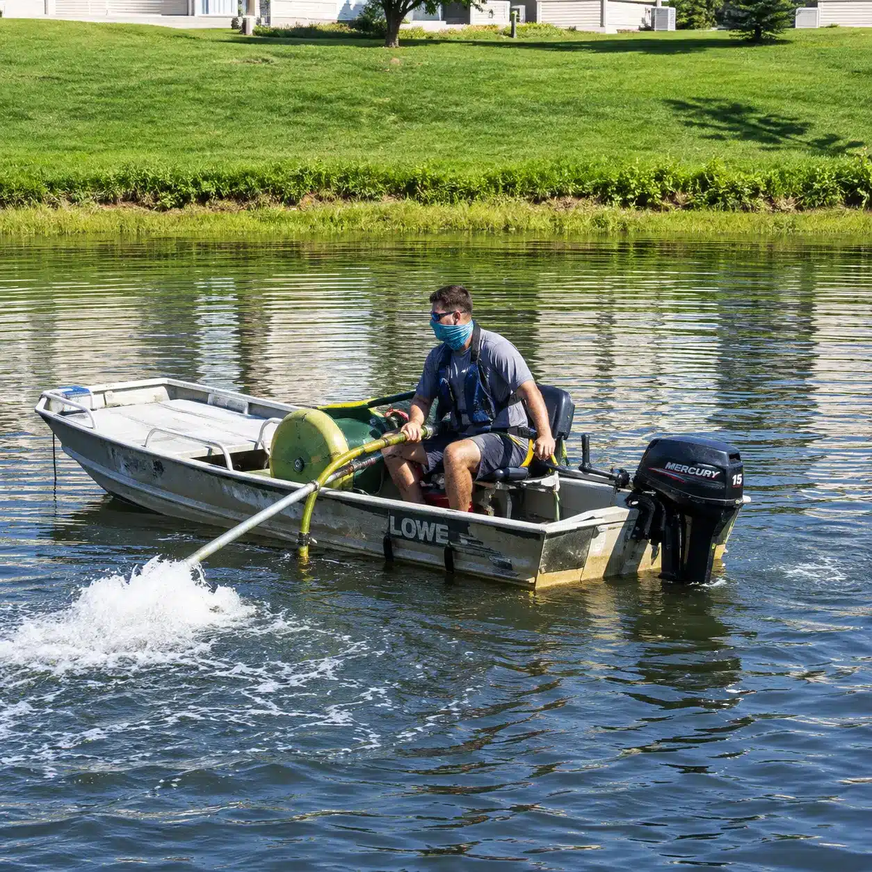 A man wearing a mask operates a small metal boat on a pond, using a hose to spray water into the lake near a grassy bank.