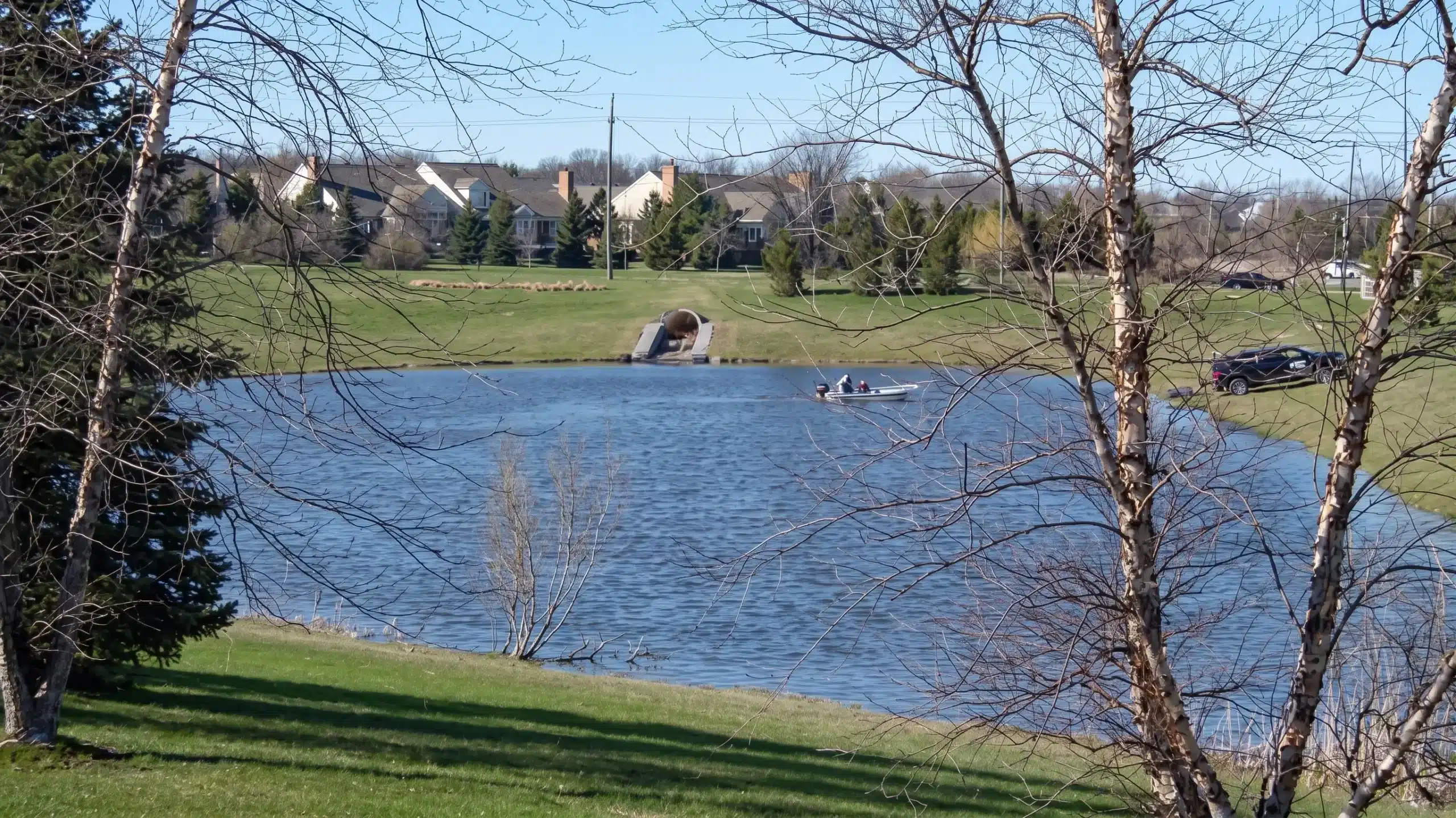 A small lake with a boat carrying people, surrounded by grass and trees, with houses and a drainage pipe in the background under a clear blue sky.