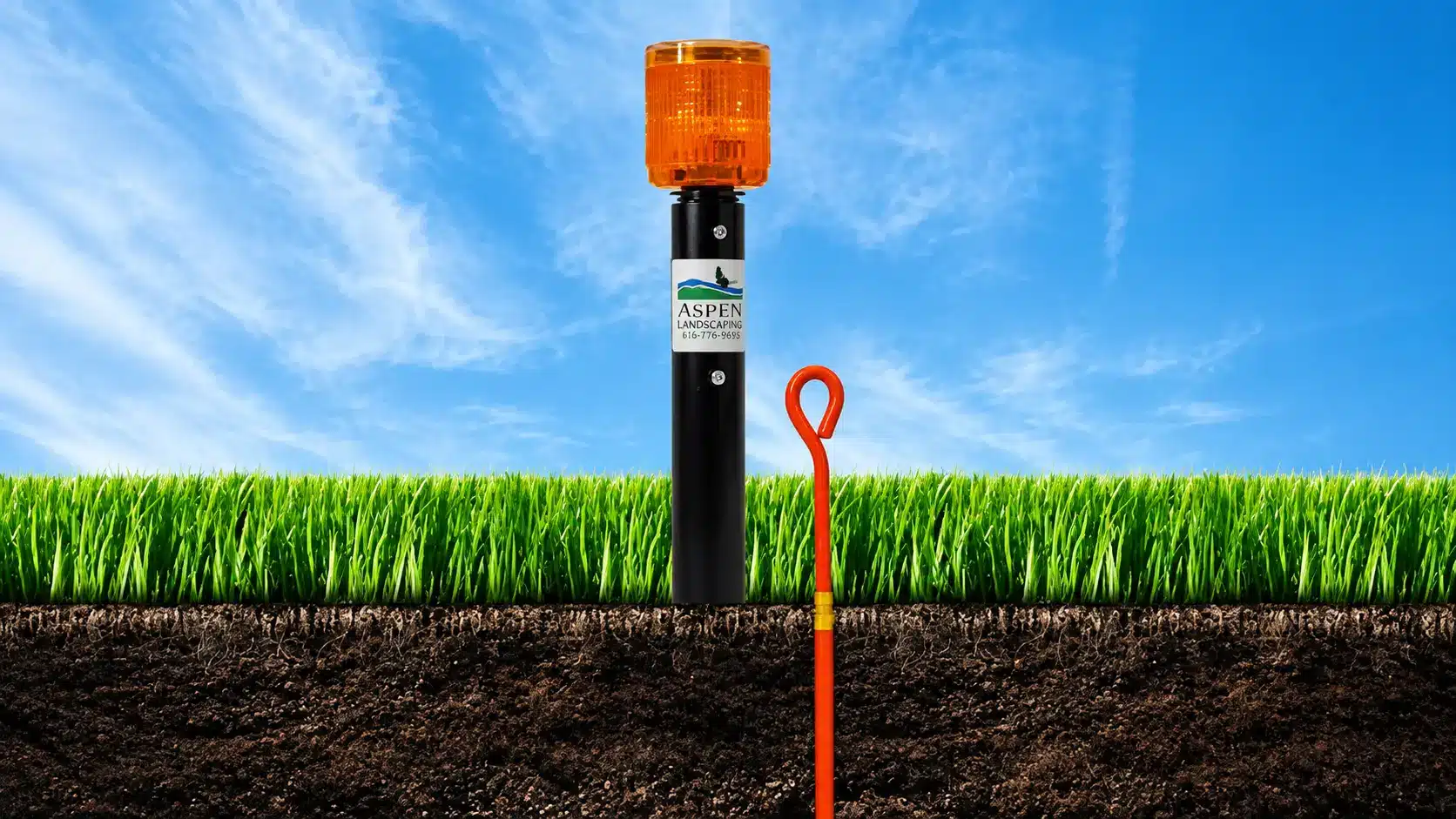 A black pole with an orange warning light and an Aspen label is installed in grass, with an orange ground marker stake next to it, against a blue sky background.