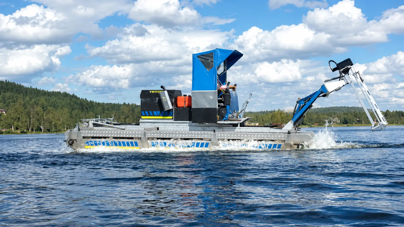 A mechanical weed harvester moves across a lake, operated by a person under a blue canopy, with forested shore visible in the background.