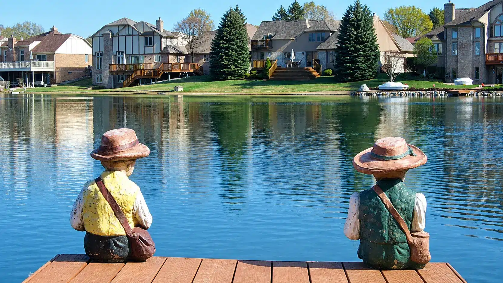 Two statue figures sit on a wooden dock facing a calm lake, with suburban houses, trees, and boats visible across the water.