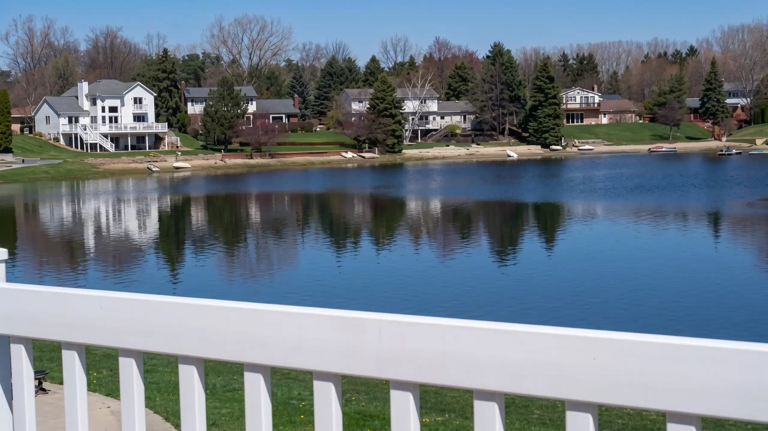 View of a peaceful lake with clear reflections of houses, trees, and boats along the shore, seen from behind a white railing under a bright blue sky.