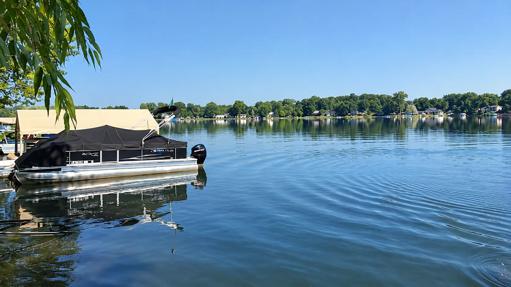 A pontoon boat with a canopy is docked on a calm lake under a clear blue sky, with trees and houses visible on the distant shoreline.